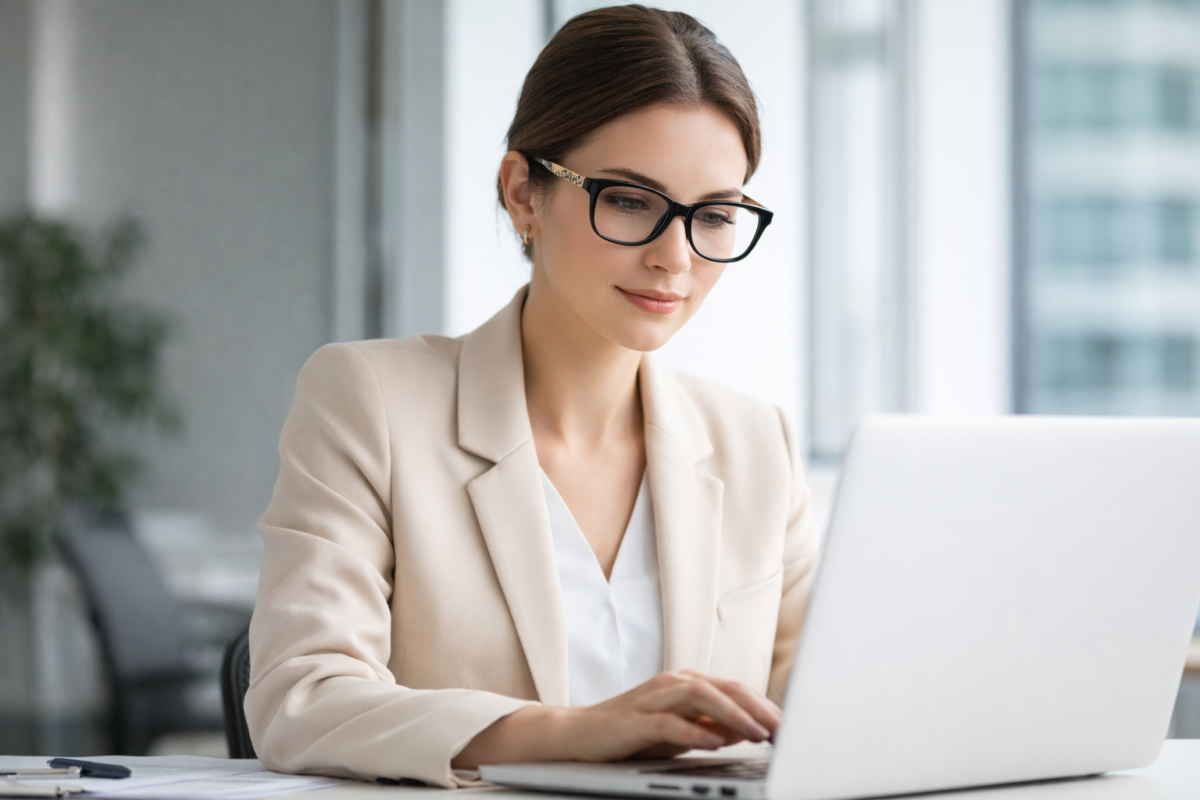 Woman wearing black square eyeglasses and a beige blazer, working on a laptop in a bright office setting.